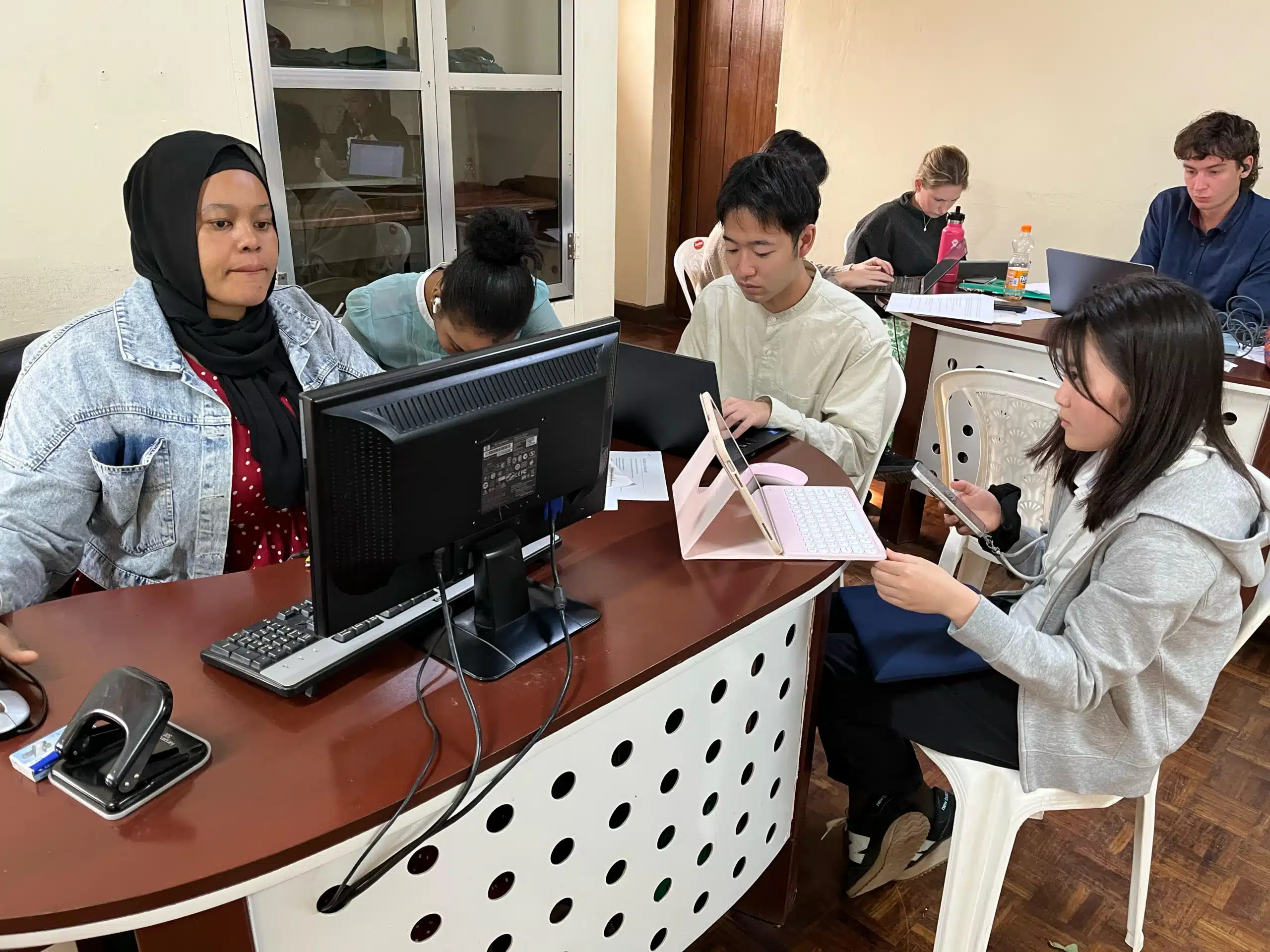 The image depicts a group of individuals engaged in a collaborative work setting, likely an educational or training environment. They are seated around a curved, modern desk with a perforated design, which accommodates multiple computer monitors.