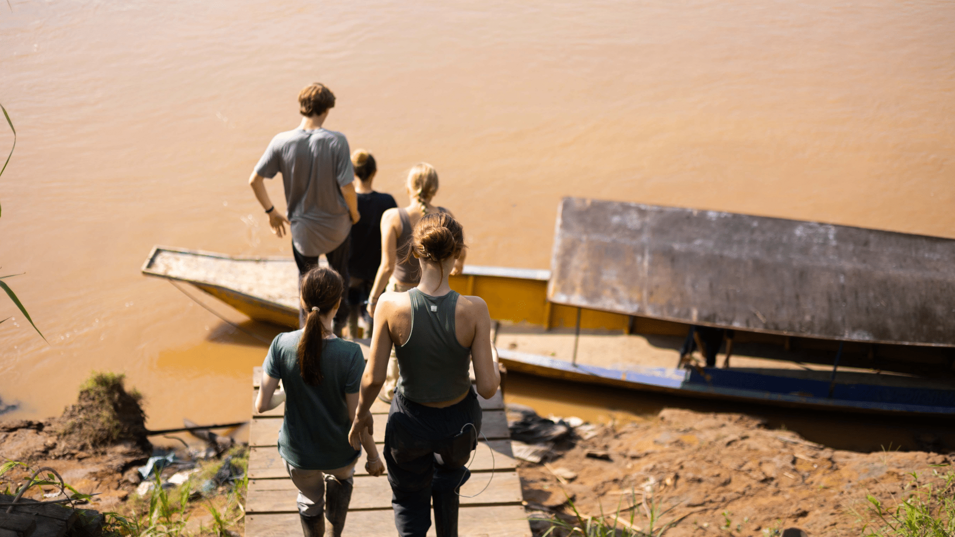 A group of young travelers seen from behind as they walk down a wooden ramp toward a long boat docked on the Amazon river in Peru.