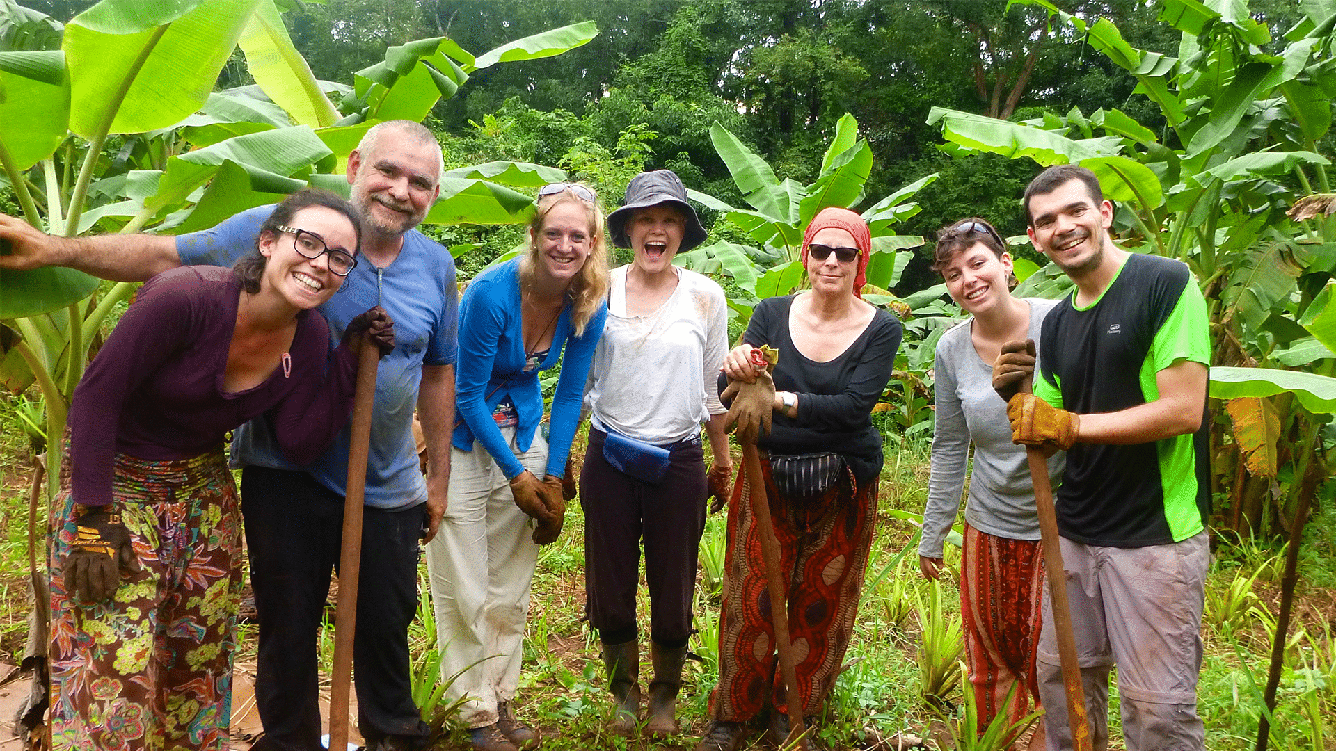 A group of seven smiling volunteers posing together in a lush, tropical field surrounded by banana trees. They are wearing casual, earthy clothing and heavy-duty work gloves, with several individuals holding tall wooden-handled tools like shovels or spades, leaning on them while smiling at the camera.