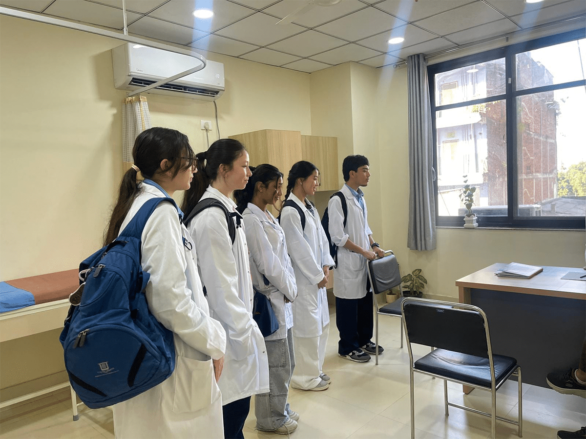 A line of five medical interns or volunteers wearing white lab coats and backpacks listening attentively inside a clinic in Nepal.