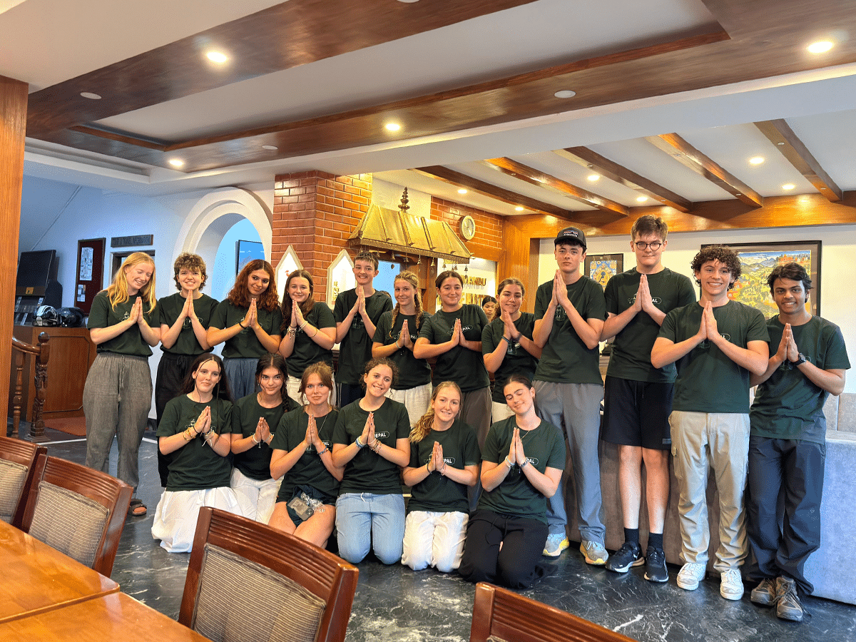 A large group of teenagers posing together indoors, with the front row kneeling and the back row standing. They are all wearing matching dark green t-shirts that say "Projects Abroad Nepal" and holding their hands together in a traditional Namaste greeting gesture.