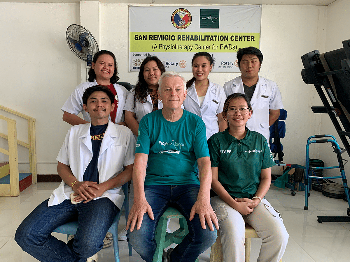 A group of seven people poses together inside the San Remigio Rehabilitation Center in the Philippines. An older man in a teal "Projects Abroad" T-shirt sits in the center foreground, with local staff and medical professionals wearing white lab coats and green staff shirts.